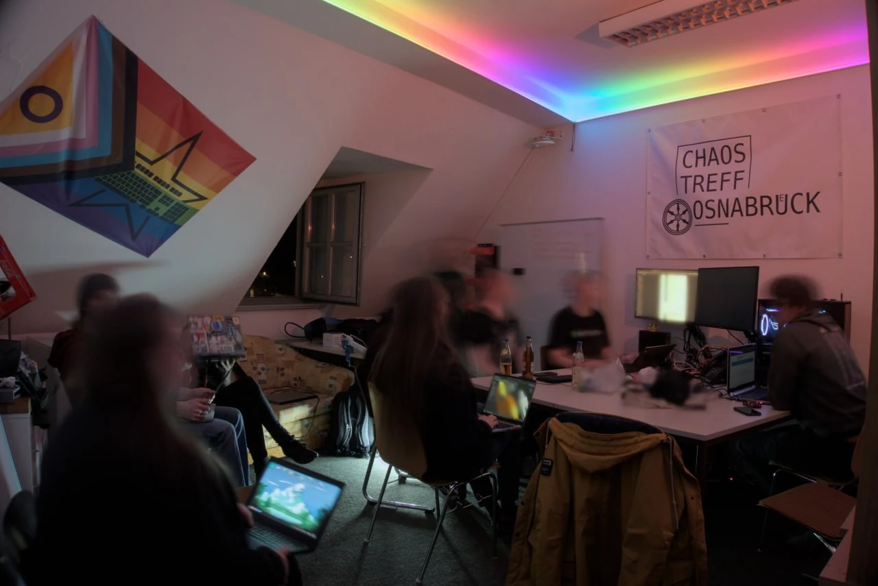 A photo of the hackspace. Humans with laptops sit around a table and on a couch. On the walls there is a banner with the Chaostreff Osnabrück logo, a progress flag and a whiteboard. The space is illuminated by a led strip with rainbow colors.